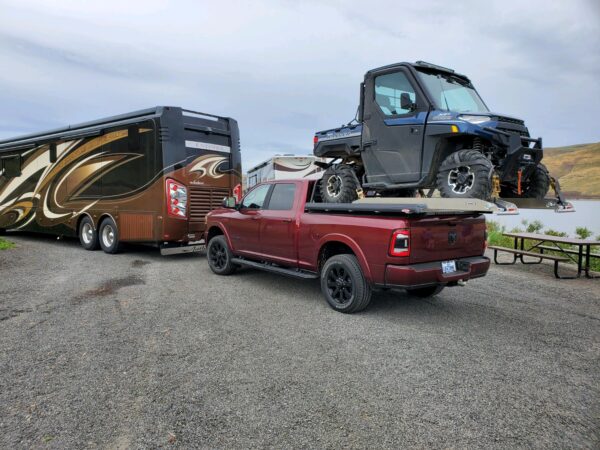 A red truck with a truckboss deck and a UTV secured to the deck. Hills and a lake are in the background with a couple motor homes.